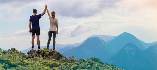 Back view of young tourist couple, athletic man and slim girl standing with raised arms holding hands on rocky mountain top enjoying fantastic panorama. Tourism, traveling and climbing concept.