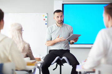 Contemporary teacher with tablet looking at one of students while voicing online data at lesson