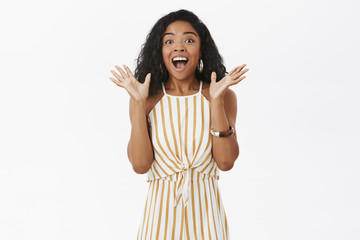 Portrait of joyful excited dark-skinned woman telling awesome news shaking raised palms in joy and happiness posing with opened from delight mouth gazing amazed at camera over gray wall