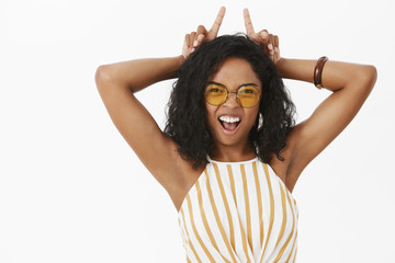Fototapeta premium Showing all sides of temper. Portrait of daring energized and confident funny young african american woman in sunglasses and striped top showing teeth holding index fingers on hear like devil horns