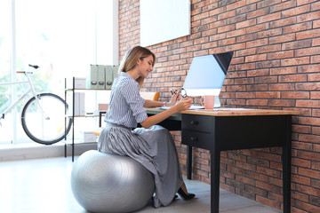 Beautiful businesswoman sitting on fitness ball at desk in office. Workplace exercises - Powered by Adobe
