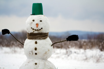 Big smiling snowman with bucket hat, scarf and gloves on white snowy field winter landscape, blurred black trees and blue sky copy space background. . Merry Christmas and happy new year greeting card. © bilanol
