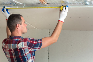 Young handsome man in casual clothing takes measurement of drywall suspended ceiling connected to...