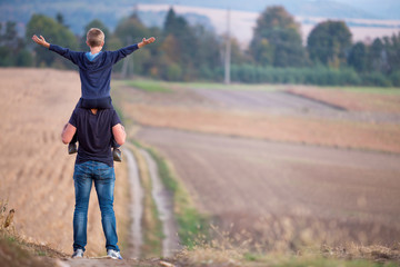 Back view of athletic father carrying on shoulders son walking through grassy field on blurred foggy green trees and blue sky background. Active lifestyle, family relations, weekend activity concept.