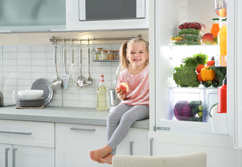 Cute little girl with apple sitting near open refrigerator in kitchen