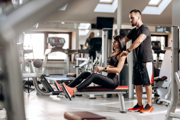 Obraz na płótnie Canvas Beautiful slim girl dressed in black sport clothes is doing exercises for the abdominals on a special exercise machine under the supervision of a coach in the gym