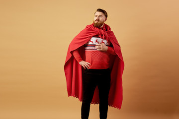 Red-haired man with beard dressed in a red and white sweater with deer and red cape   and sleepers stands on a beige background