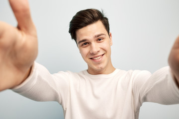 Funny dark-haired guy dressed in a white long sleeve t-shirt taking selfie on a white background
