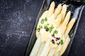Traditional boiled white asparagus with sauce hollandaise and herbs as top view on a plate with copy space left