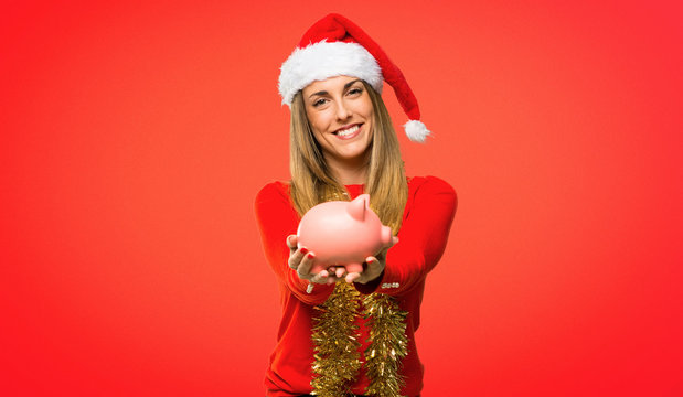 Blonde woman dressed up for christmas holidays taking a piggy bank and happy because it is full on red background
