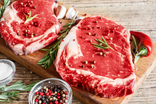 Raw beef rib eye steaks with spices and rosemary on wooden board over rustic wooden background