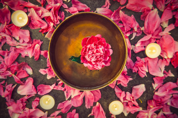 Top view tibetan singing bowl with floating inside in water pink peony flower. Burning candles and petals on the black stone background. Meditation and Relax. Exotic massage. Flatlay. Selective focus.