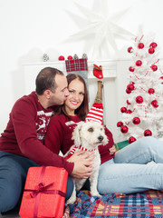 Happy couple with dog, all in Christmas clothes sitting near Christmas tree