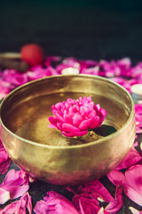 Tibetan singing bowl with floating in water pink peony flower. Burning candles and petals on the black stone background. Meditation and Relax. Exotic massage. Vertical. Selective focus. Copy space.