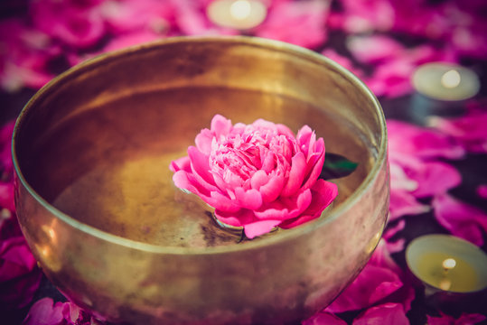 Tibetan singing bowl with floating inside in water pink peony flower. Burning candles and petals on the black stone background. Meditation and Relax. Exotic massage. Selective focus.