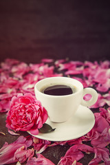 Close up cup of freshly brewed coffee and pink peony flower on the saucer on black background with petals. Good morning concept. Vintage toning. Vertical card. Soft selective focus, copy space.