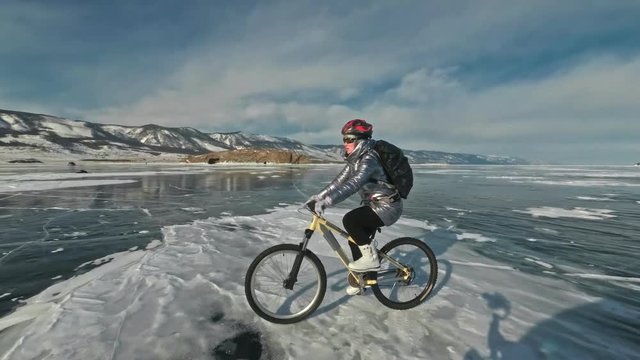 Woman is riding bicycle on the ice. The girl is dressed in a silvery down jacket, cycling backpack and helmet. Ice of the frozen Lake Baikal. The tires on the bicycle are covered with special spikes