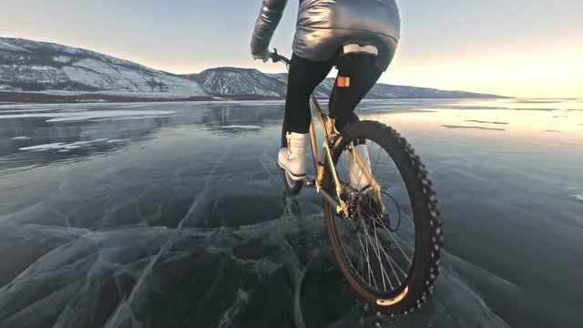 Woman is riding bicycle on the ice. The girl is dressed in a silvery down jacket, cycling backpack and helmet. Ice of the frozen Lake Baikal. The tires on the bicycle are covered with special spikes