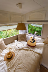 Inside the camper van. Unfilled bed, pillows, guitar, book, hat, white wooden decoration of the house on wheels.