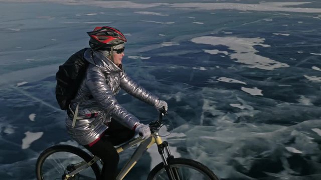 Woman is riding bicycle on the ice. The girl is dressed in a silvery down jacket, cycling backpack and helmet. Shooting with a quadrocopter drone. The tires on the bicycle are covered with special