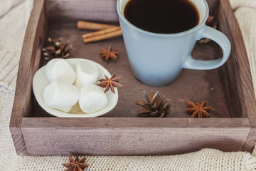 Cup of coffee on rustic wooden tray, sweet marshmallow and warm woolen sweater. Cozy autumn or winter weekend or holidays at home. Fall home decoration with hot drink mug. Hygge morning style concept