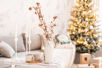vase with cotton branches. Light living room with Christmas tree. Comfortable sofa, high large Windows