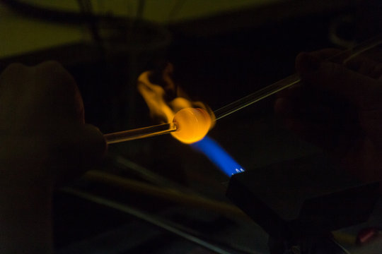 A crafts person is using a blow torch to melt and form a piece of glass. The flame is blue and golden where it is hitting the glass. 