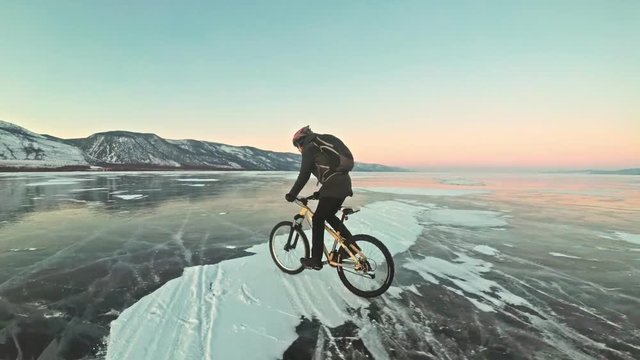 Man is riding a bicycle on ice. The cyclist is dressed in a gray down jacket, backpack and helmet. Ice of the frozen Lake Baikal. The tires on the bicycle are covered with special spikes. The traveler