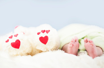 Fototapeta na wymiar small tender legs of the baby, together with the paws of a Teddy bear with hearts on a white blanket