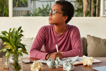 Thoughtful black girl in eyewear focused aside, tries to gather with thoughts, writes notes in blank sheet of paper, surrounded with notepad, notes ideas for startup, modern smart phone on table