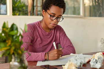 Successful designer with black skin, boyish haircut, writes down plan for week on blank paper, dressed in casual turtleneck sweater, spectacles, thinks of new ideas for drawing, develops project