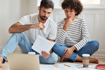 Obraz premium Indoor shot of serious mixed race woman and man study private arrangement, work from home, drink coffee, sit crossed legs on floor, read paper content attentively, try arrive at common conclusion