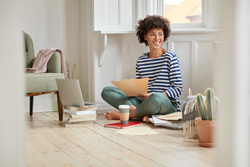 Cheerful black woman feels pleased, poses in lotus pose, reads financial report, enjoys calm domestic atmosphere, surrounded with modern laptop computer, develops new strategy, learns information