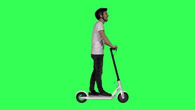 Happy young man riding an electric scooter, wearing a helmet, waves hello and looks around over a green screen.