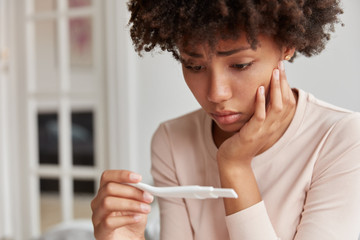 Displeased frustrated African American woman looks stressfully at pregnancy test, finds out she will become mother, looks desperately at result. Close up shot of young lady has problems with fertility - obrazy, fototapety, plakaty