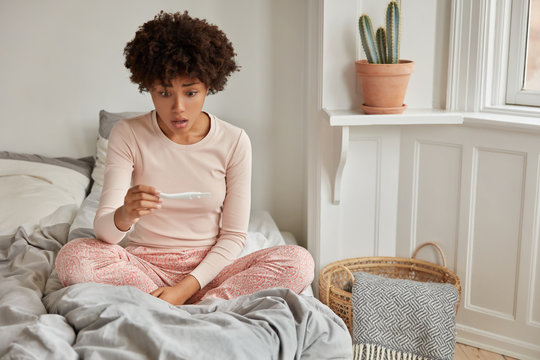 Unwanted child. Shocked dark skinned young woman looks with puzzlement, sits crossed legs on bed, stares at positive result of pregnancy test, has distracted facial expression, models in bedroom