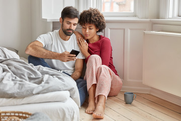 Relaxed multiethnic boyfriend and girlfriend sit on floor at home, wear casual domestic clothes, read new post on internet website, use smart phone and high speed internet, drink hot beverage