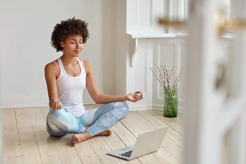 Tuinposter Calm lady with Afro hairstyle, dressed in sportswear, meditates on floor in empty room, listens spiritual practices lessons on laptop computer, poses in lotus pose, tries to relax. Yoga concept © Wayhome Studio