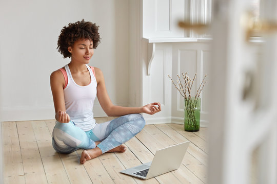 Calm lady with Afro hairstyle, dressed in sportswear, meditates on floor in empty room, listens spiritual practices lessons on laptop computer, poses in lotus pose, tries to relax. Yoga concept