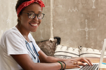 Photo of smiling fashionable lady has white teeth, wears casual clothes, keyboards on notebook...