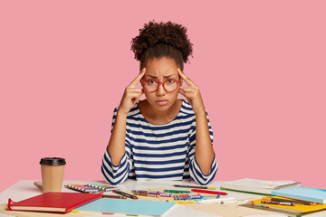 Dissatisfied dark skinned woman keeps index fingers on temples, gathers with thoughts, dressed in striped clothes, surrounded with crayons and blank sheet of papers. People and work concept.