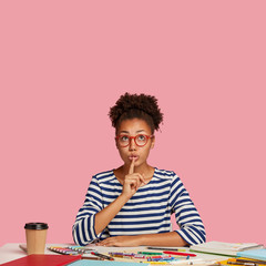 Vertical indoor shot of secret woman dressed in casual striped clothes, makes hush gesture, sits at...