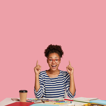 Astonished overjoyed black woman with Afro hair combed in bun, wears transparent glasses, points with both index finger upwards, shows free space for your advertising content against pink wall