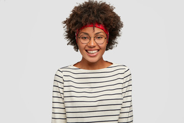Happiness concept. Beautiful black woman with Afro haircut, smiles gladfully at camera, dressed in striped clothing, models against white background, being in high spirit as rejoices promotion at work