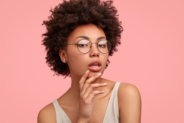 Headshot of self confident dark skinned young female keeps hand on chin, raises head with pride, shows her natural beauty, has Afro hairstyle, wears optical glasses, isolated over pink background