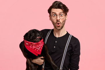 Surprised unshaven man in black shirt with braces, holds small pedigree dog with bandana, being shocked to have conversation with vet, finds out about pets disease, poses against pink background - obrazy, fototapety, plakaty