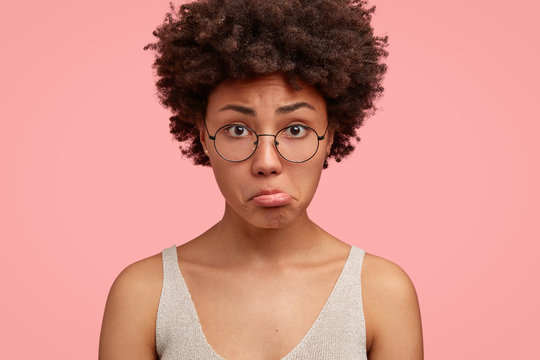Cropped close up shot of displeased black woman purses lower lip, looks in bewilderment, has sad expression, bushy hair, dressed in casual vest, models against pink background. Facial expressions