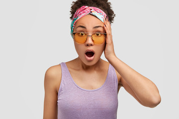 Portrait of shocked beautiful woman with dark skin, Afro hair, wears colourful headband, shocked by astonishing news, dressed in light purple vest, poses against white studio wall. Human emotions