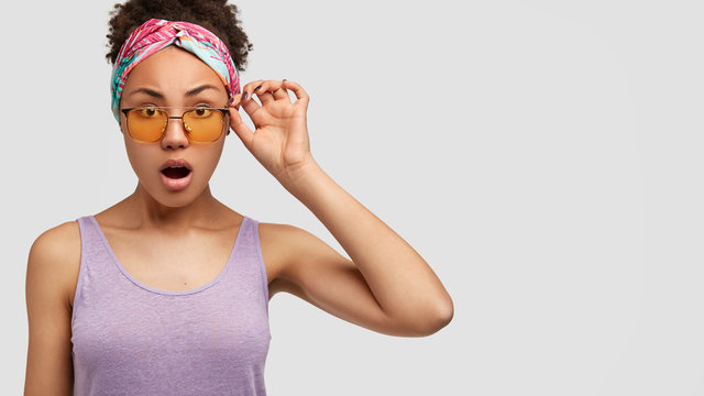 Image of beautiful mixed race female model shocked by bad news, wears headband and casual vest, keeps hand on rim of yellow trendy sunglasses, models against white background with free space