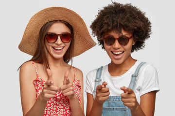 Positive women of different race, make gun gesture directly at camera, indicate with fore fingers, wear summer clothes, choose you to travel with them and having holiday trip. You, its time for rest